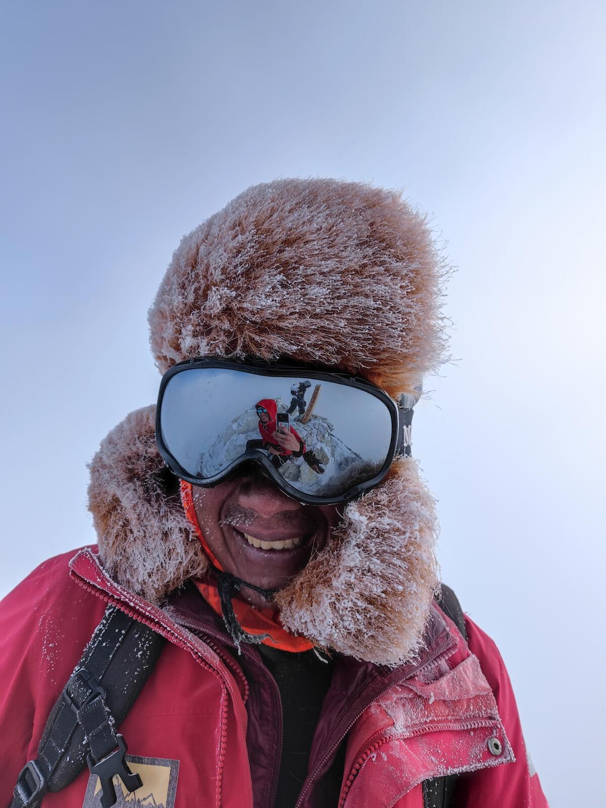 A frost-covered fur hat, with the summit scene reflected in ski goggles.